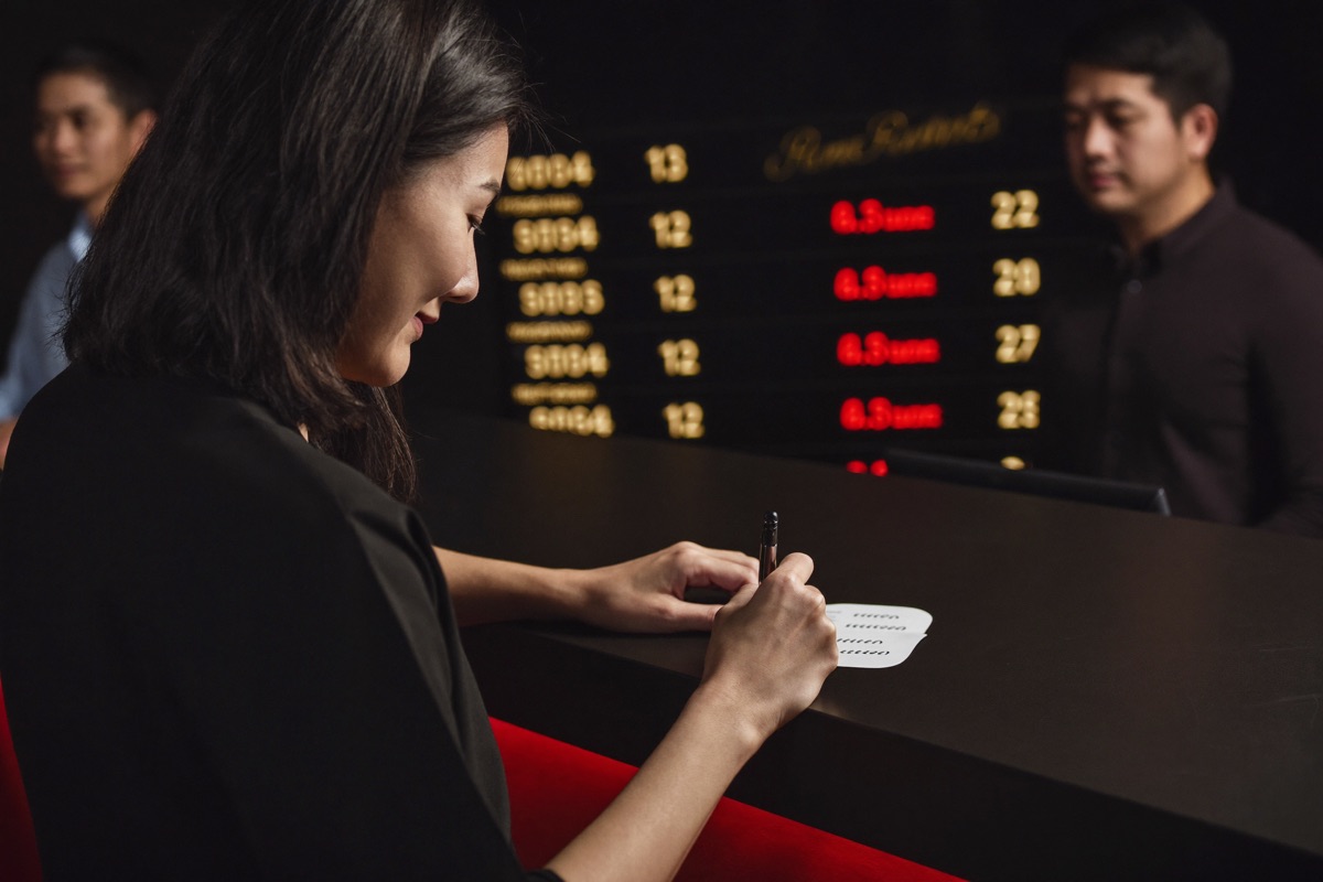 A numbers counter under a gold board of drawn digits, a woman handing a slip across a near black counter to a clerk who is stamping it