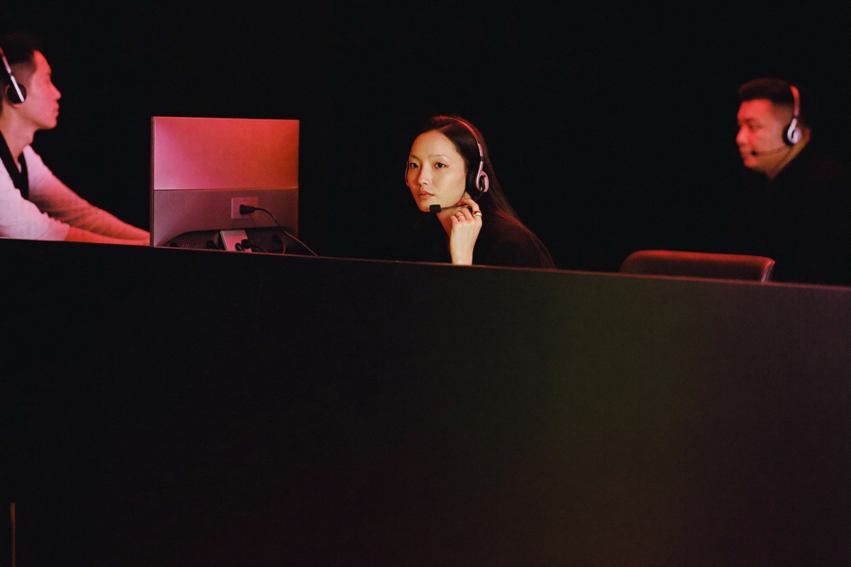 A support desk with three headset staff at near black terminals, one of them turning to speak to a visitor leaning on the crimson counter