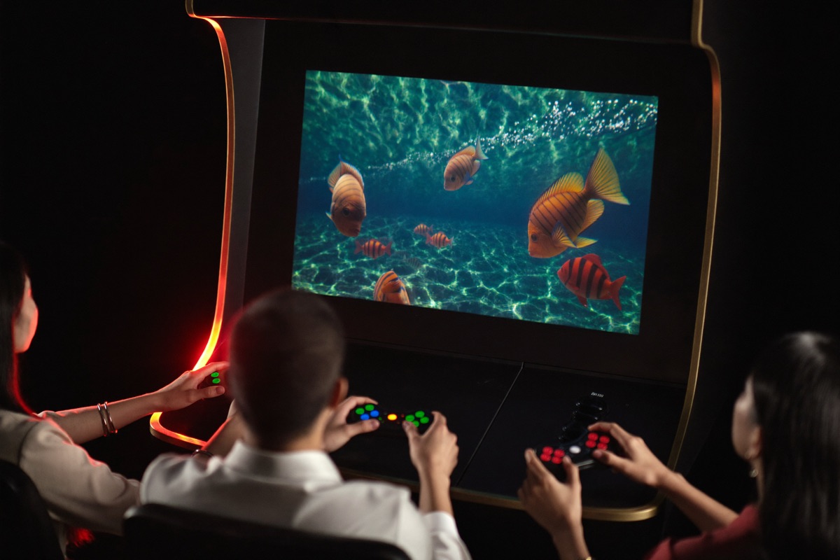 Two friends at a wide low cabinet with a blue green underwater screen, each holding a mounted gun grip, gold coin counters climbing along the top edge of the screen