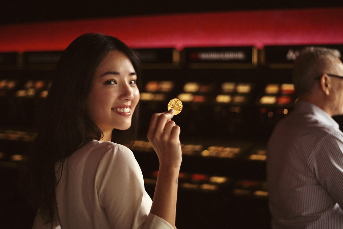 A young couple sharing one stool at a gold trimmed cabinet, the woman pointing at a small credit panel on the screen while the man holds a single note