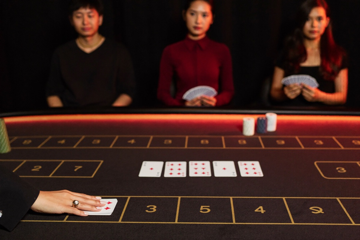 A live baccarat pit from the player's side, the dealer in black sliding a card off the shoe while three seated players watch, gold chips stacked on crimson felt