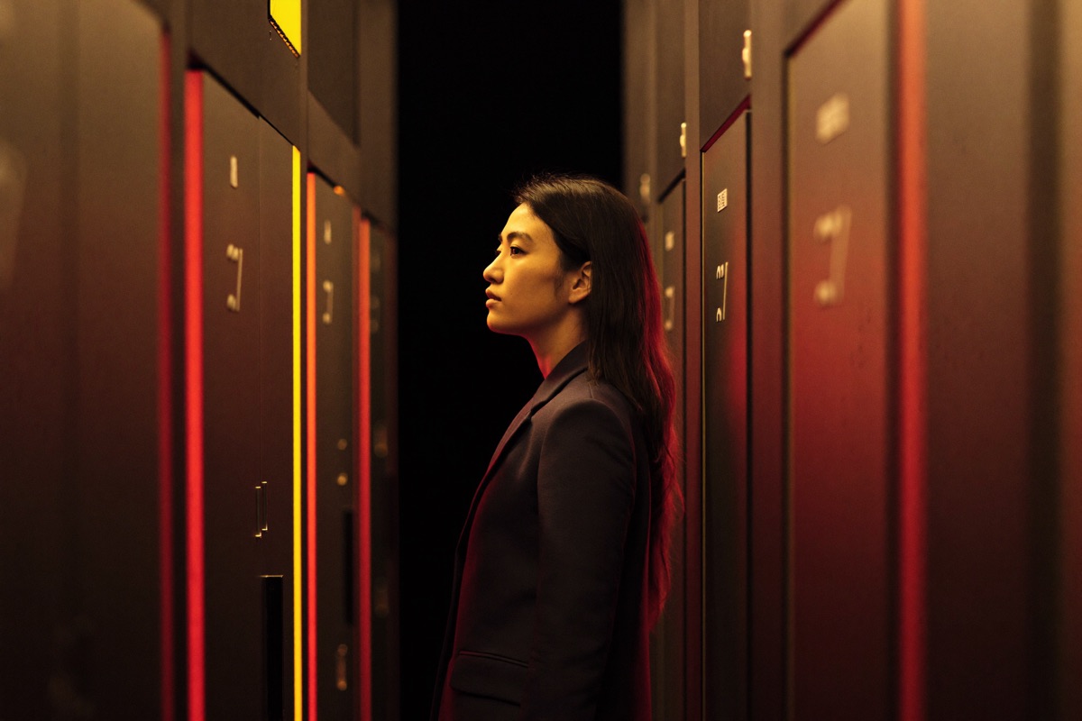 A wide casino floor at night from the walkway, gold lit slot cabinets down the left and crimson felt baccarat tables down the right, a woman in a crimson jacket leaning over the nearest cabinet to press spin while two friends watch from behind her