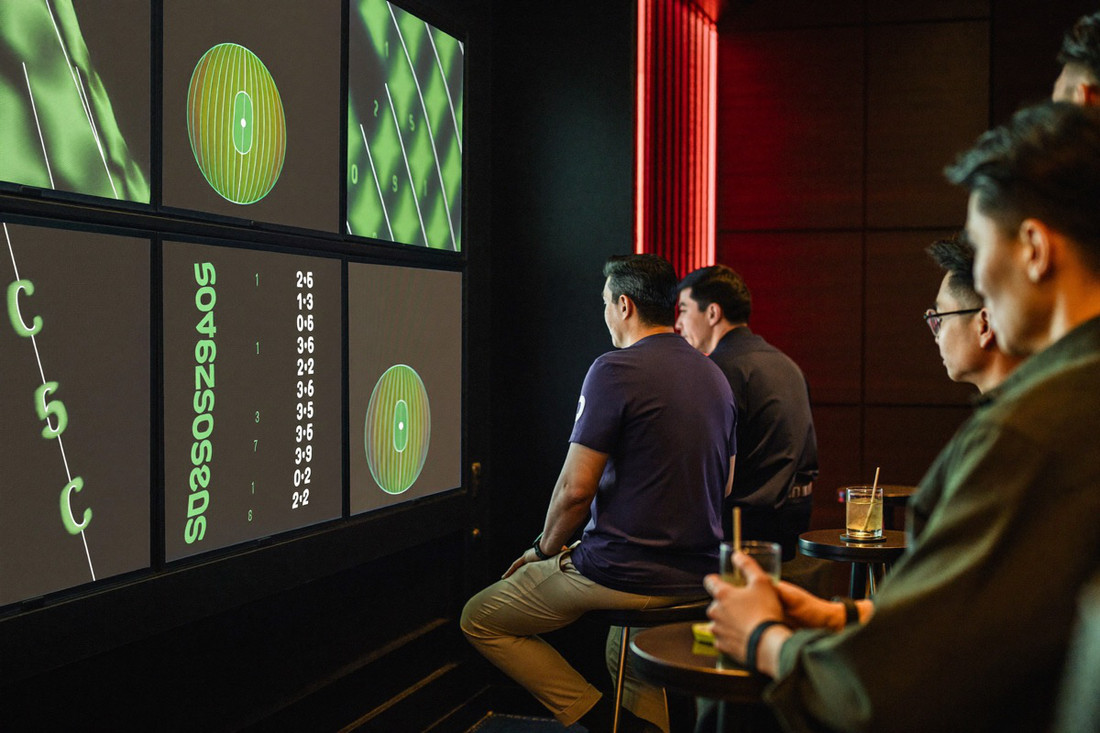 A sports room with a wall of screens showing a football match, two men standing at a crimson counter comparing a printed slip against the gold odds board above them