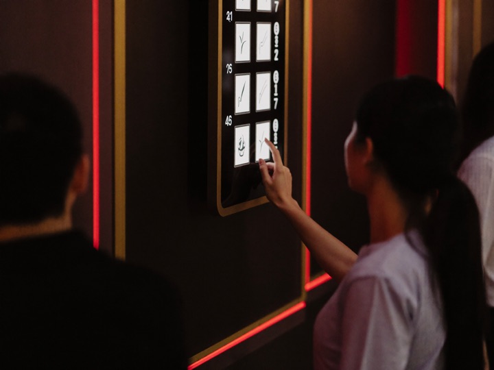 A row of gold lit cabinets with a crimson card reader glowing on the middle one, a woman tapping her phone against it while a host watches from the aisle